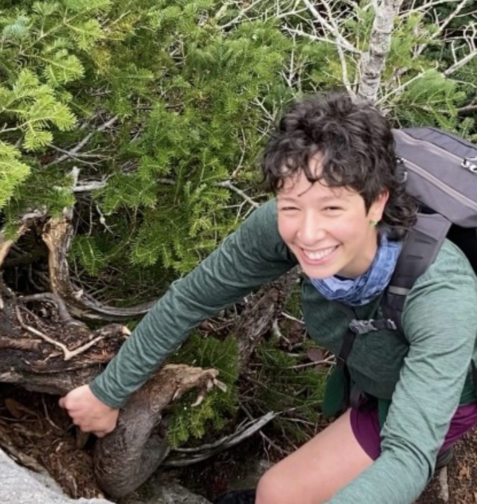 Person smiling while climbing rocks, surrounded by greenery, wearing a backpack and green shirt.