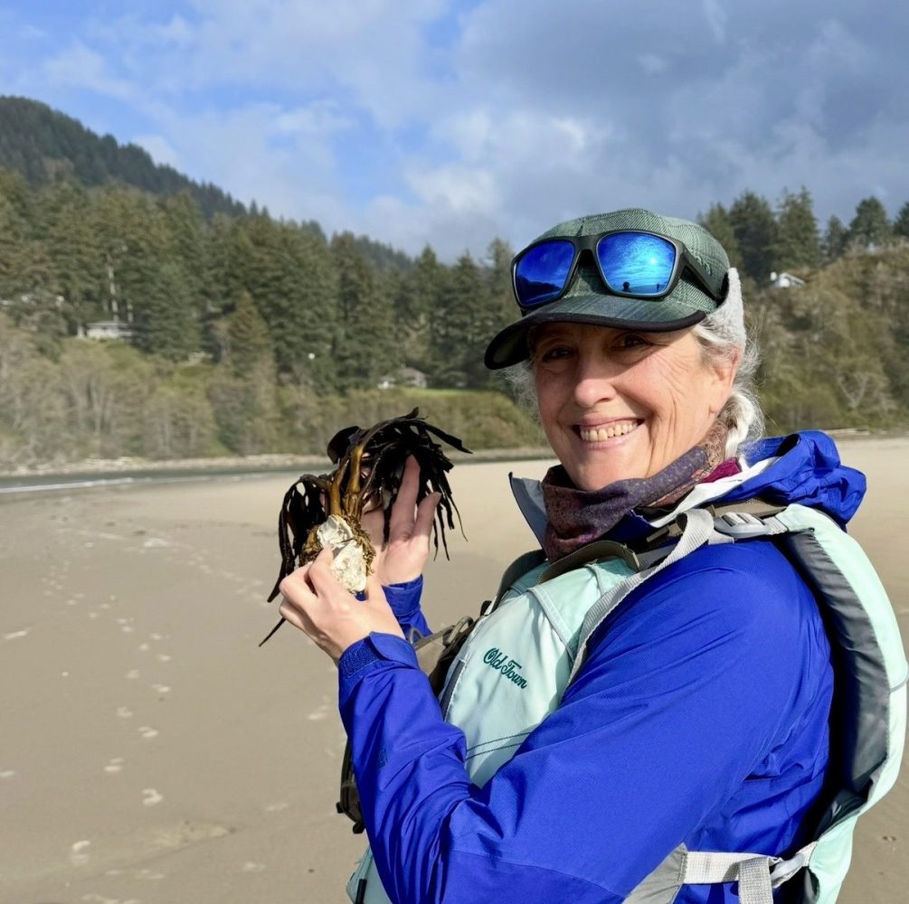 Person in blue jacket smiles, holding seaweed on a sandy beach with trees in the background.