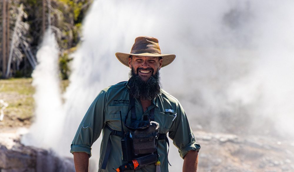Man in hat with camera gear smiles near a geyser.