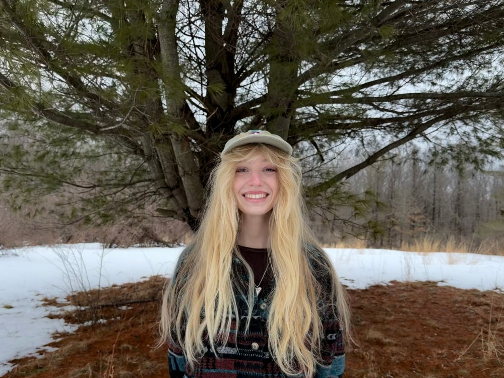 Person with long hair and cap smiling in front of a tree with snow-covered ground.