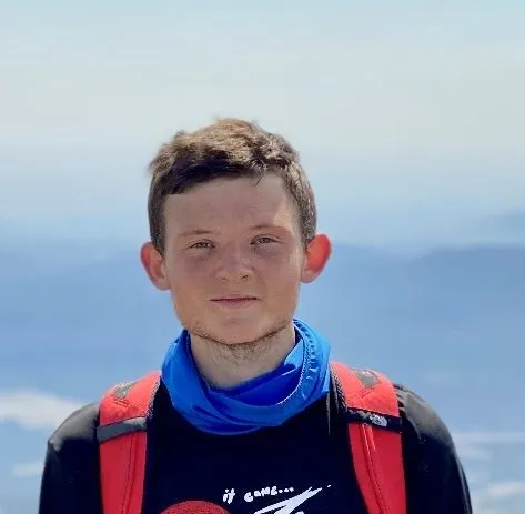 Person in black shirt with red backpack standing on mountain with blue sky and distant hills.
