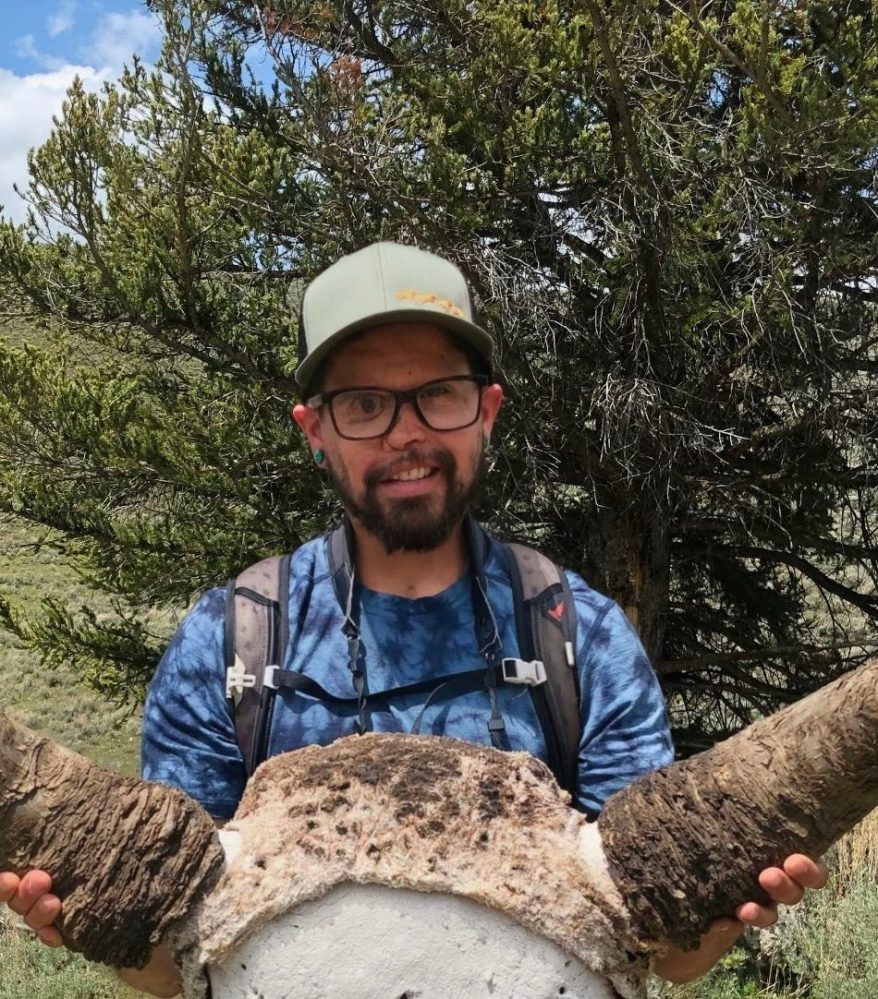 Man holding large animal skull with horns outdoors, trees in background.