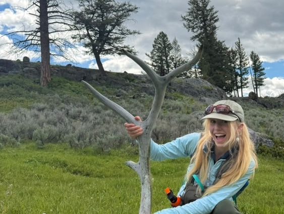 Person in outdoor gear holding a large antler in grassy area with cloudy sky.