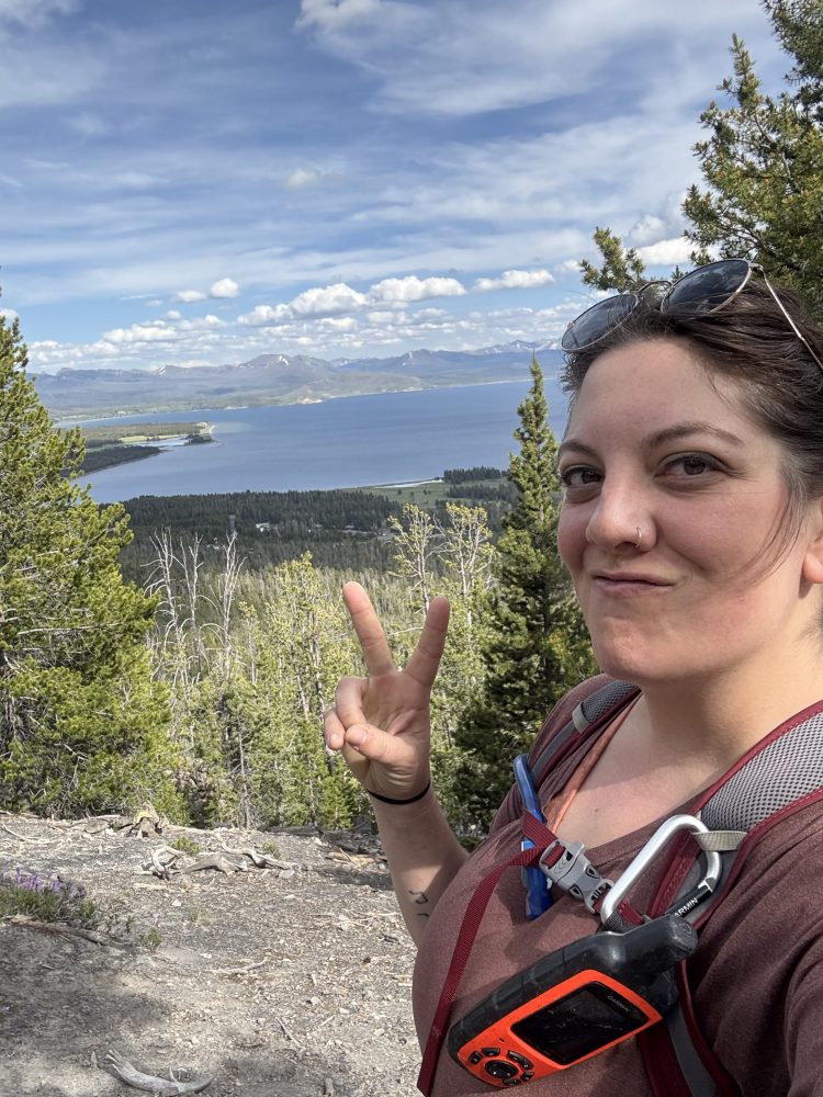 Person hiking on a trail with a scenic view of a lake, mountains, and trees, making a peace sign.