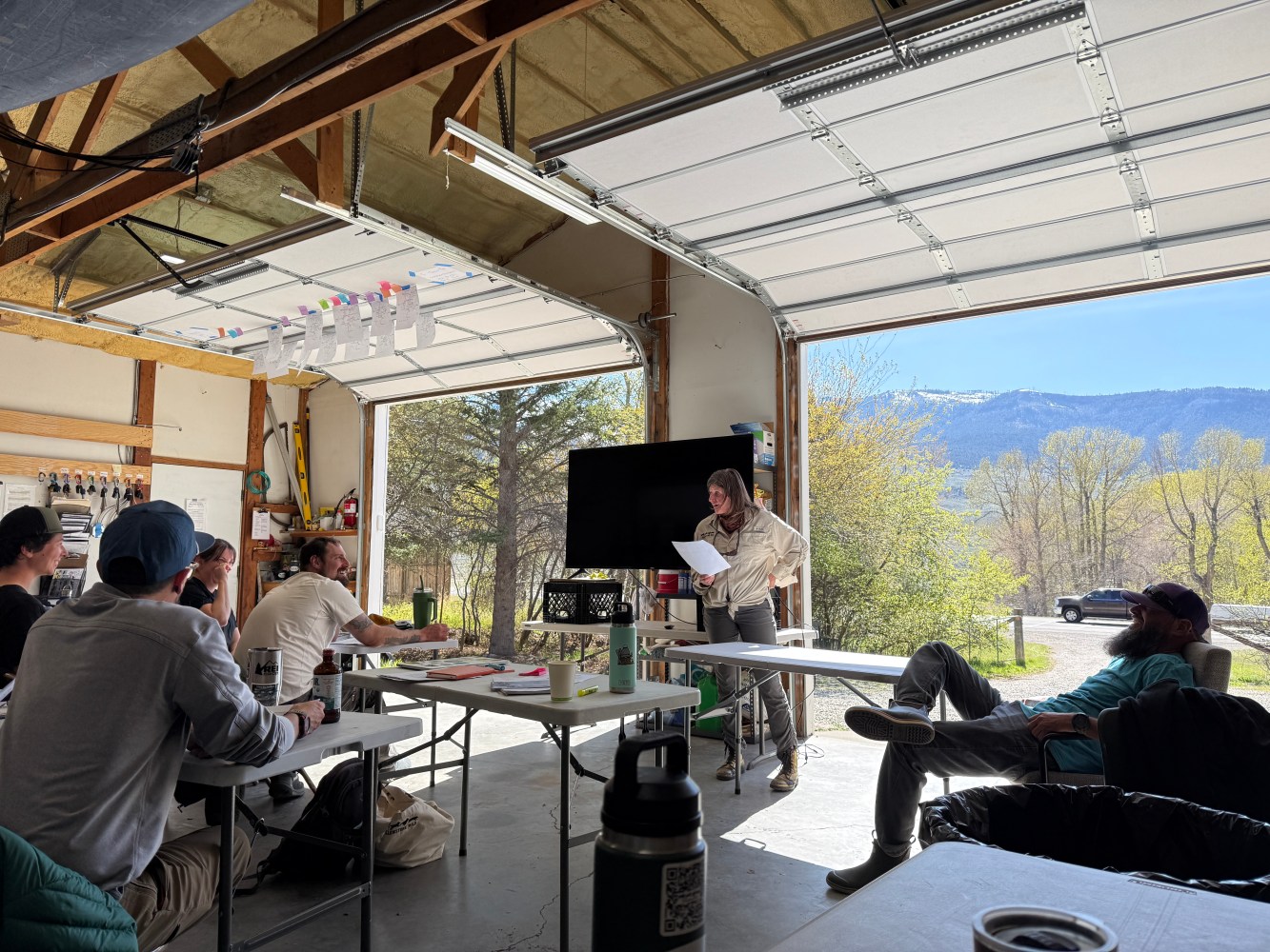 People in a garage-like room, one standing near a screen, with a mountain view outside.