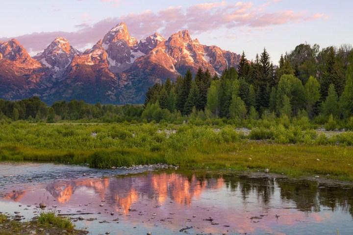 Mountain range at sunset with trees, grass, and reflective water in foreground.