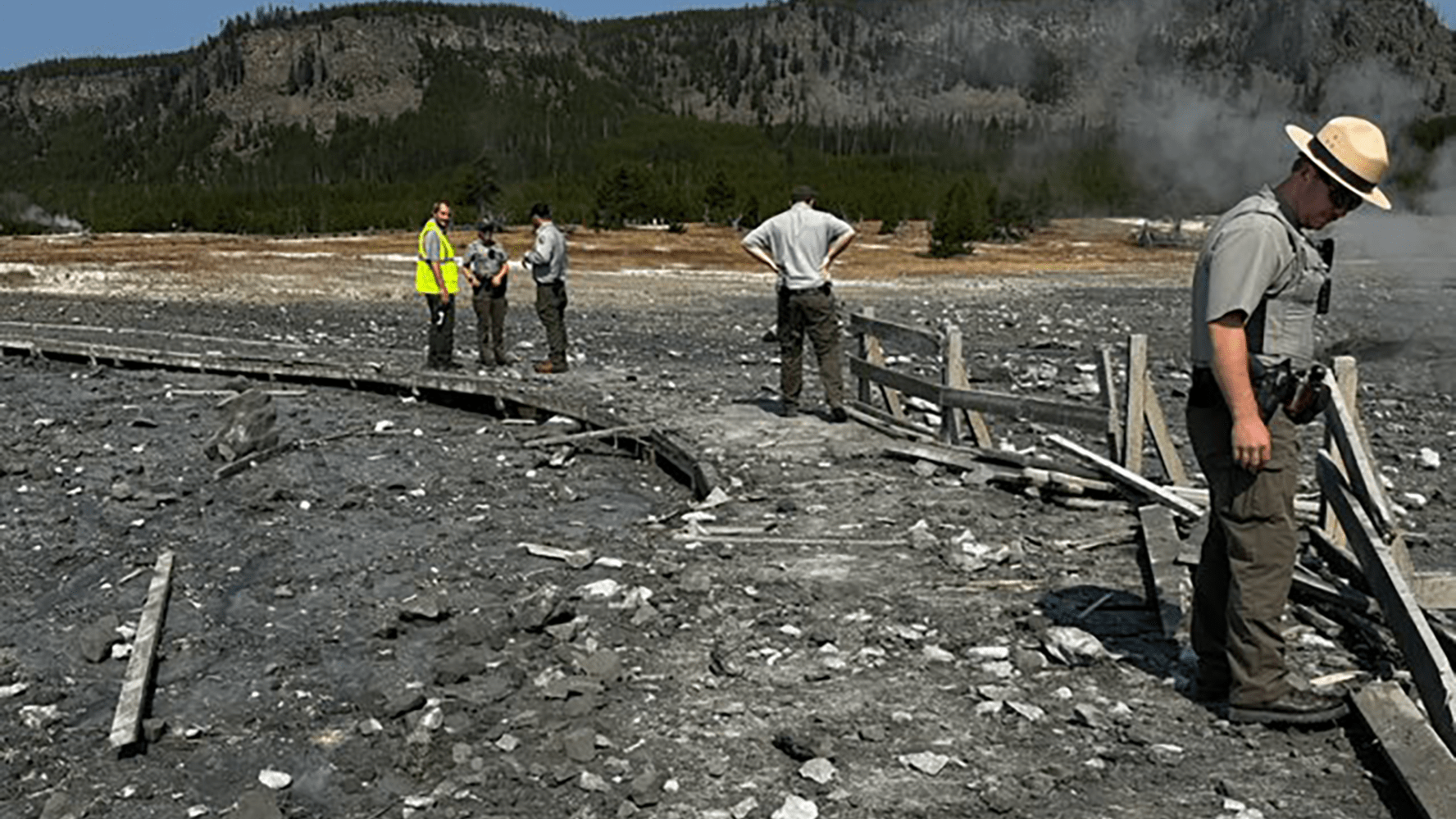 People in a geothermal area, some wearing uniforms, with mist and damaged walkway.