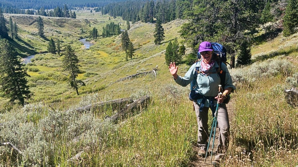 Hiker with backpack waves on a trail in a scenic valley with trees and a river.