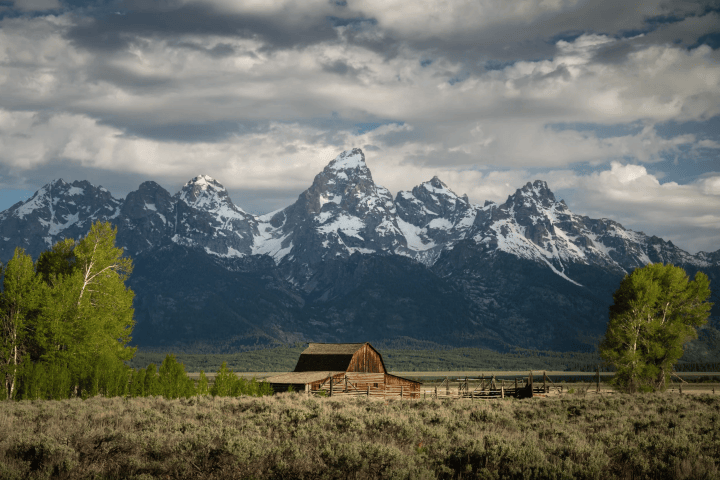 Wooden barn with snow-capped mountains in background under cloudy sky.